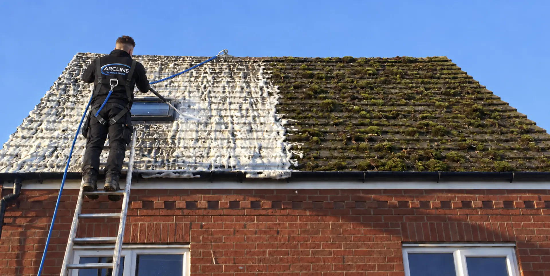 Arcline on a ladder spraying treatment across a mossy roof, half the roof treated and half still covered in moss